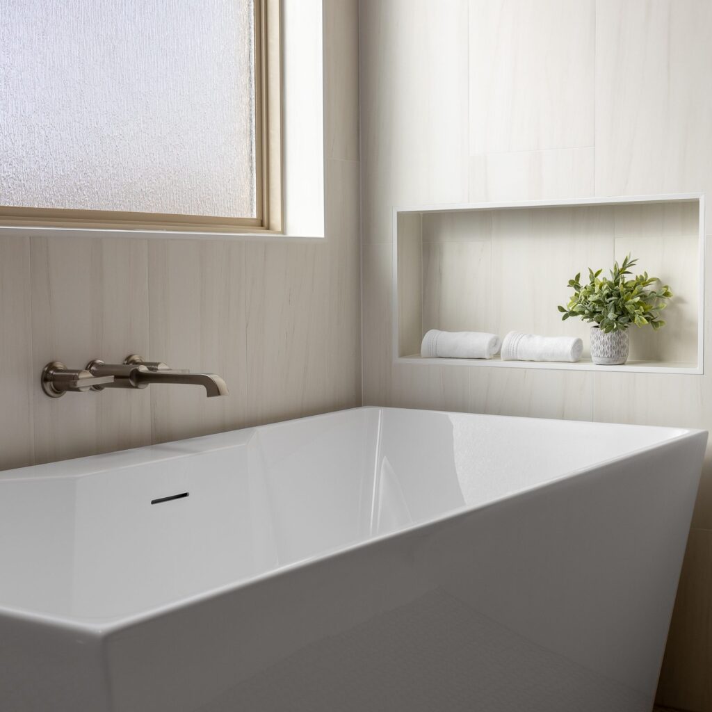 Freestanding soaking tub with wall-mounted faucet and lit niche in a primary bath remodel